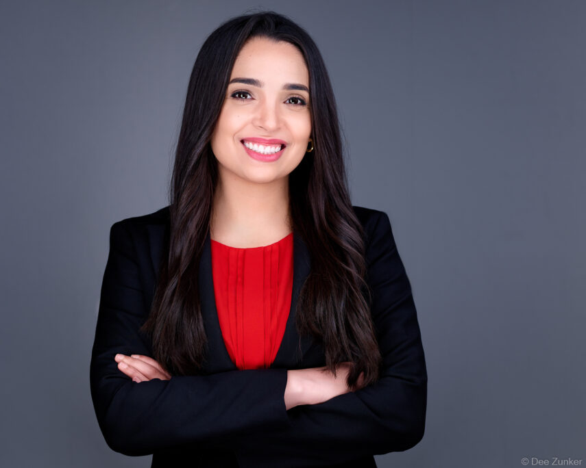 Female attorney in black blazer and red top poses with arms crossed for a professional studio headshot in Houston, gray background.
