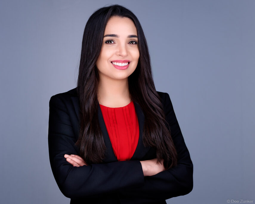 Professional corporate headshot of a woman in black blazer and red top, arms crossed, smiling, studio gray background, Houston.