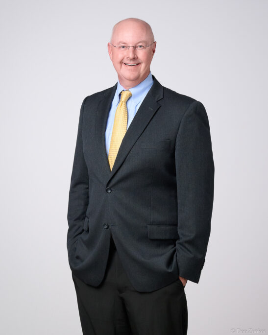 Professional corporate headshot of a smiling male financial advisor in a dark suit and gold tie, studio portrait with gray background, Houston TX.