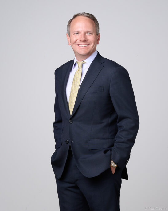Male financial executive in navy suit and gold tie poses for a corporate studio headshot in Houston, gray background.