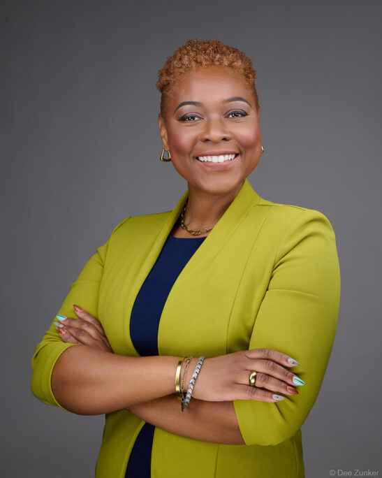 Professional corporate headshot of a smiling woman in a chartreuse blazer with arms crossed against a gray studio background, Houston TX.