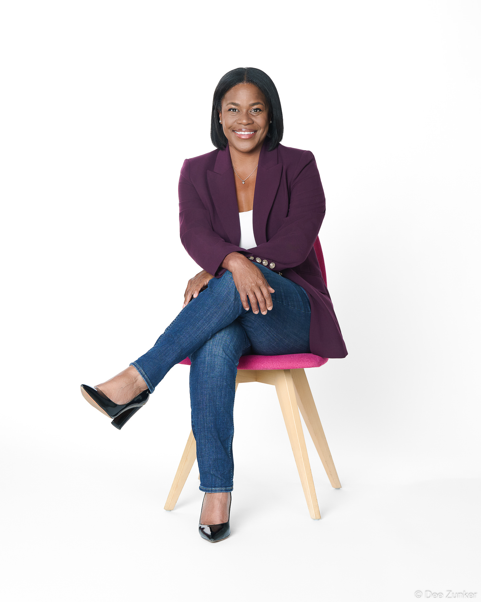 Smiling professional woman in purple blazer and jeans seated on modern chair, studio white background, Houston corporate headshot portrait.