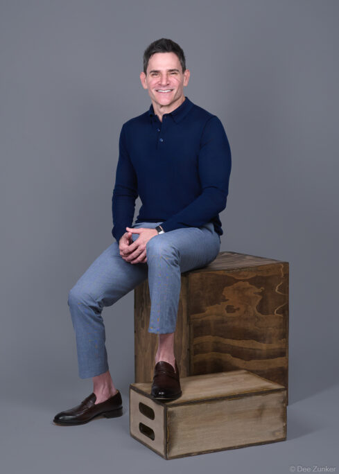 Male executive poses on wooden crate in navy polo and gray trousers, studio corporate headshot with gray background, Houston TX.