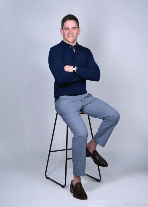 Male executive poses on stool with arms crossed in a professional Houston studio portrait against a gray background, wearing navy and gray.
