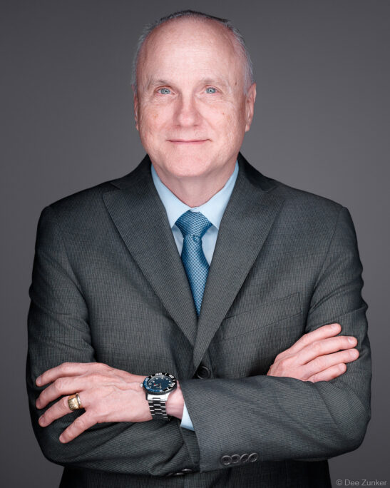 Senior male attorney in dark suit and blue tie, arms crossed, studio portrait on gray background, Houston corporate headshot photography.