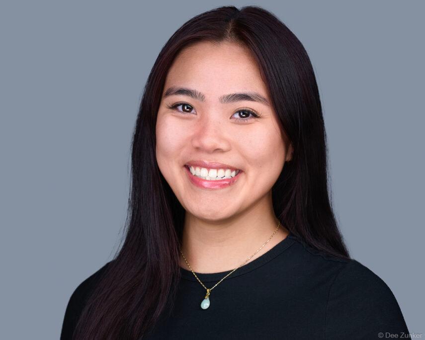 Young female architect smiles in a studio headshot against a gray background, wearing a black top and jade pendant necklace, Houston.