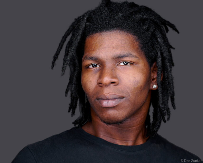 Young professional man with dreadlocks and stud earring poses for a corporate studio headshot against a gray background in Austin.