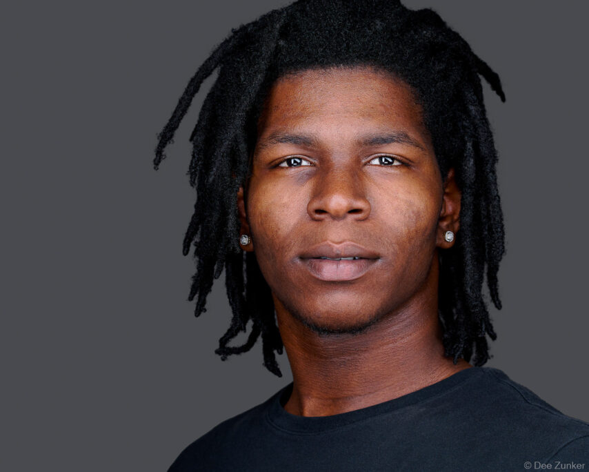 Young Black professional man with locs and stud earrings in a studio headshot against a gray background, Austin Texas.