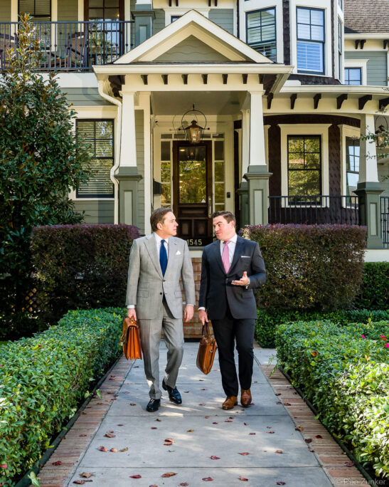 Two attorneys in business suits carrying leather briefcases walk along a pathway outside an elegant Heights Houston home