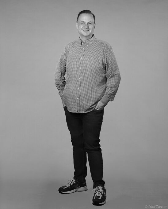 Black and white full-length studio portrait of a smiling professional man in a checkered shirt and dark jeans, gray background, Houston.