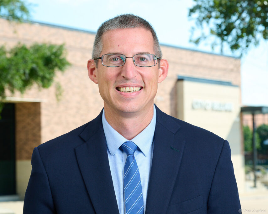 Smiling male executive in navy suit and blue striped tie, environmental corporate headshot outdoors at Bellaire city building, Houston TX.