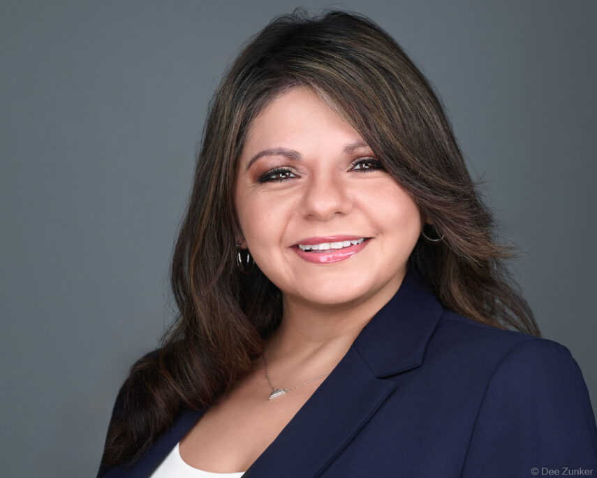 Professional corporate headshot of a female attorney in a navy blazer, smiling against a gray studio background, Houston TX.