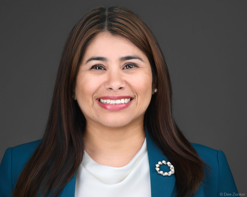 Professional corporate headshot of a smiling female attorney in a teal blazer with pearl brooch, photographed against a gray studio background in Houston.