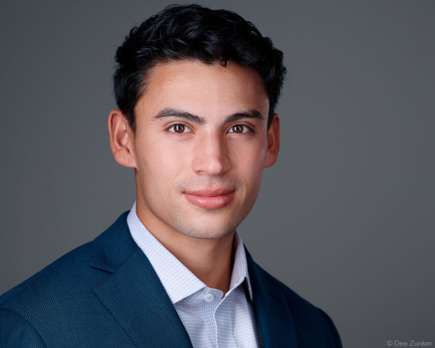 Professional corporate headshot of a young male attorney in a navy blazer against a gray studio background, Houston, TX.
