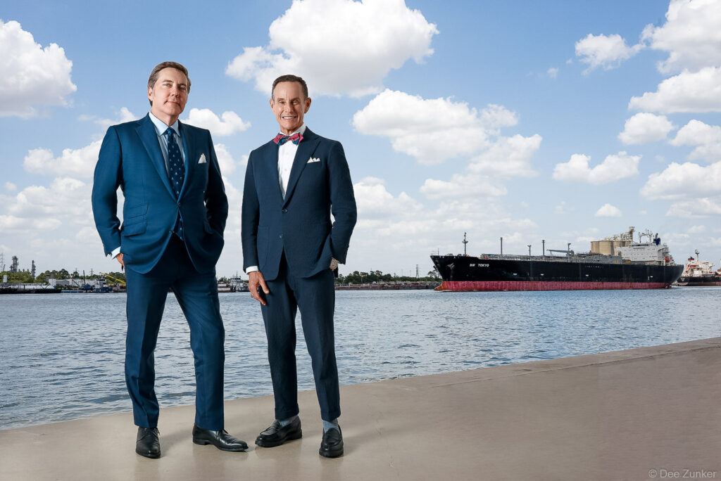 Two professional men in navy suits pose waterside at the Houston Ship Channel with a large tanker ship behind them.