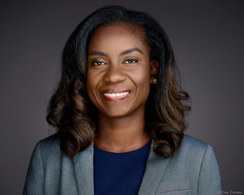 Professional woman in gray blazer smiles in studio headshot against dark gray background, corporate portrait by Houston photographer.