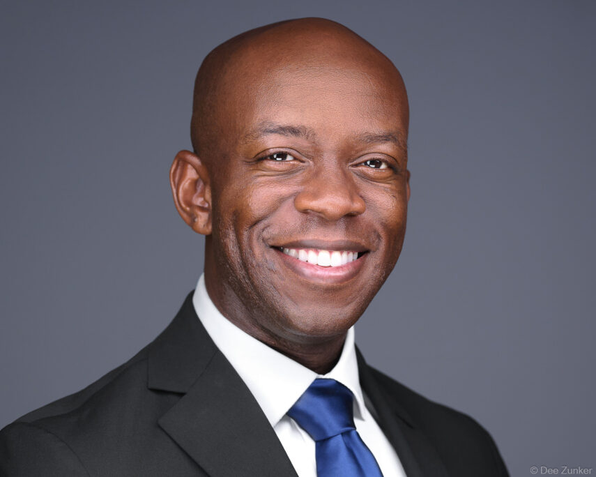Smiling bald Black male executive in dark suit and blue tie, professional studio headshot on gray background, Houston corporate photography.