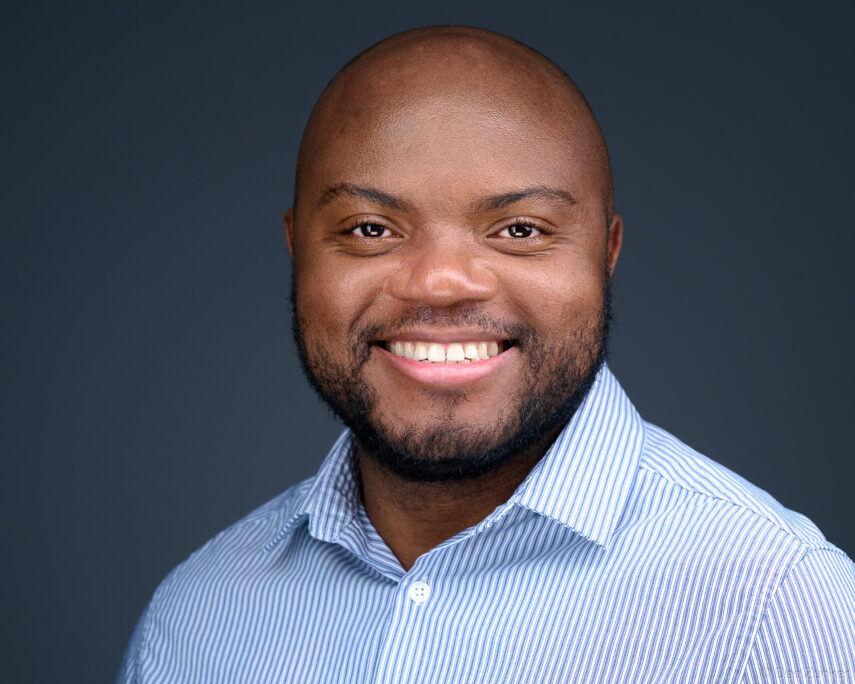 Professional corporate headshot of a smiling bald man in a blue striped shirt against a dark gray studio background, Houston TX.