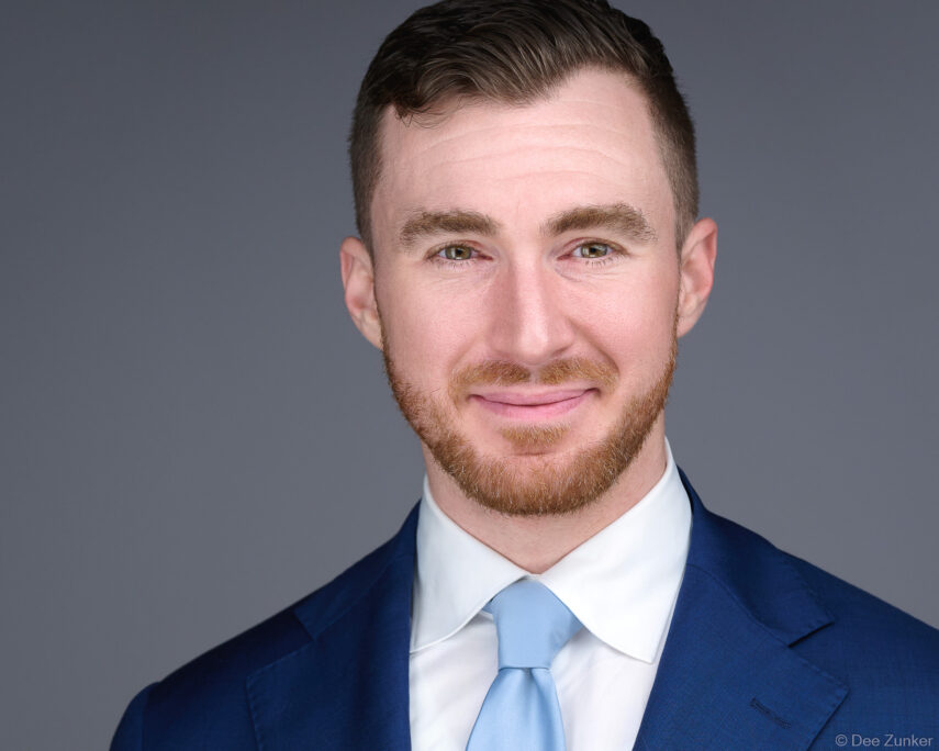 Professional corporate headshot of a man with a red beard wearing a navy suit and light blue tie against a gray studio background in Houston.