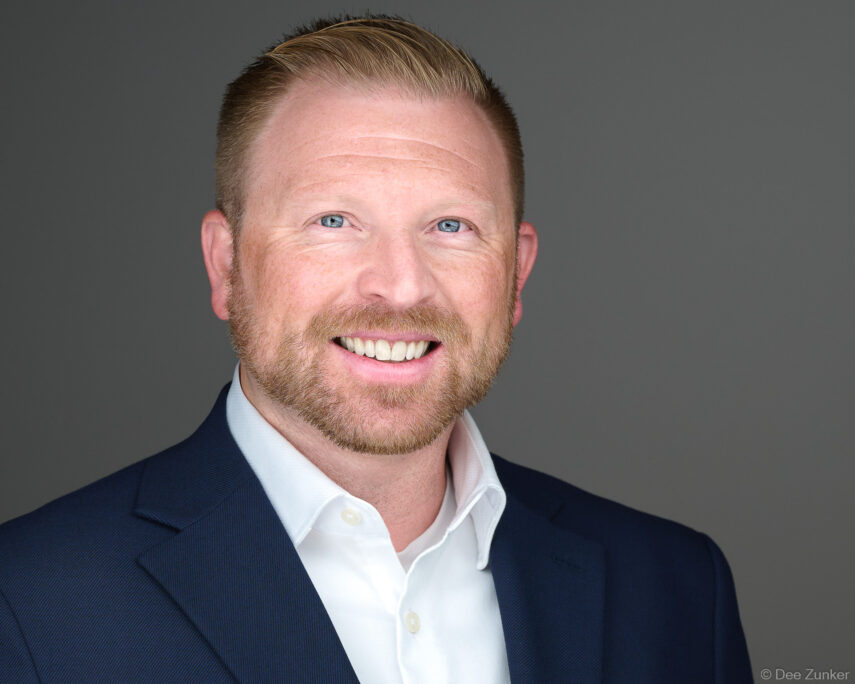 Professional corporate headshot of a smiling male executive with blue eyes and red beard, wearing a navy blazer, shot against a gray studio background in Houston.