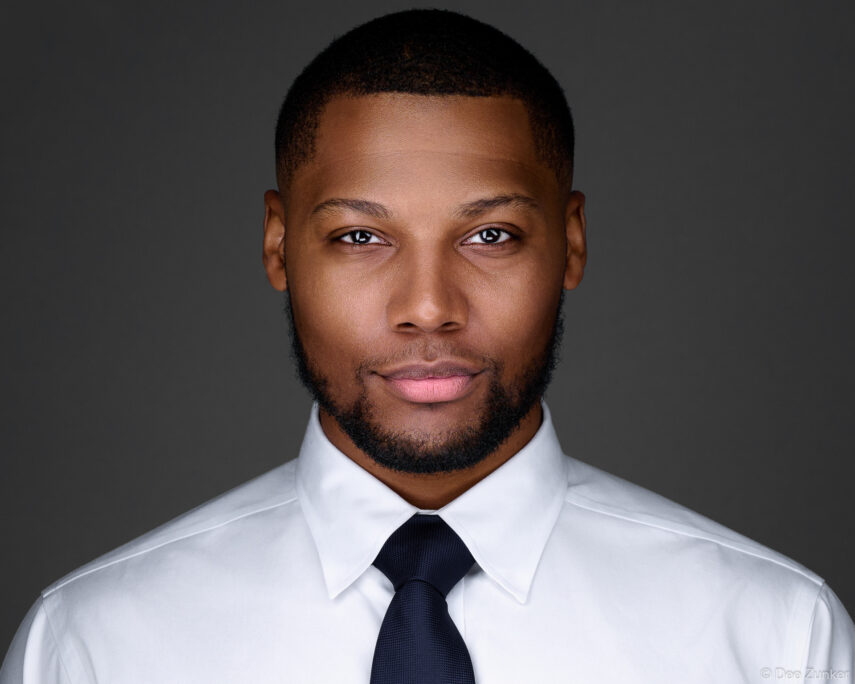 Professional corporate headshot of a man in a white dress shirt and navy tie against a gray studio background, Houston TX