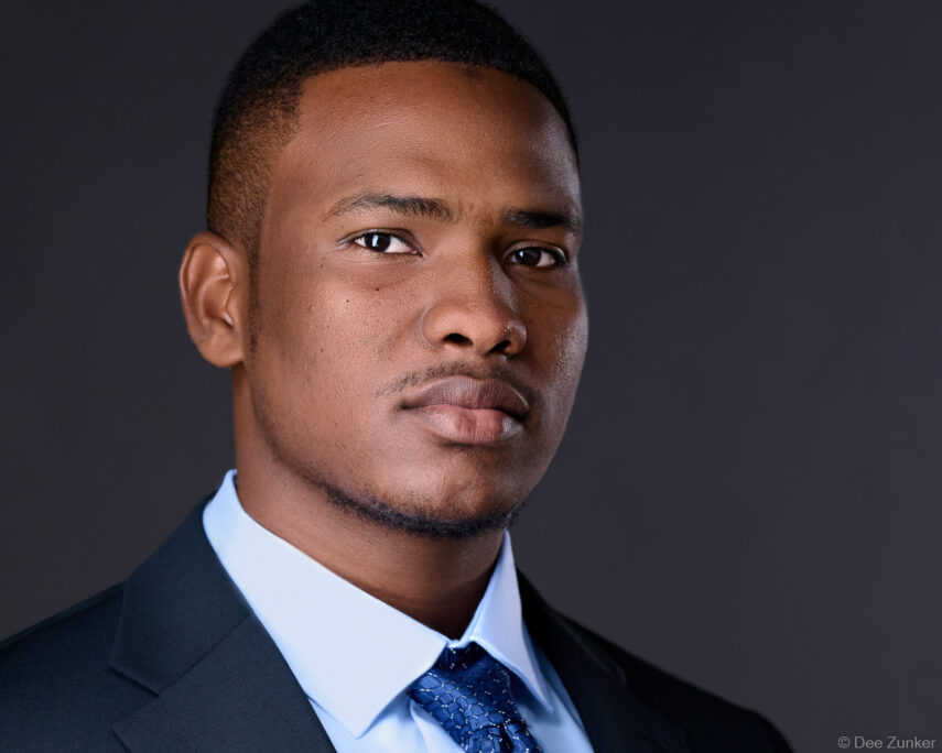 Professional corporate headshot of a young male attorney or consultant in a dark suit and blue tie against a gray studio background, Houston TX.