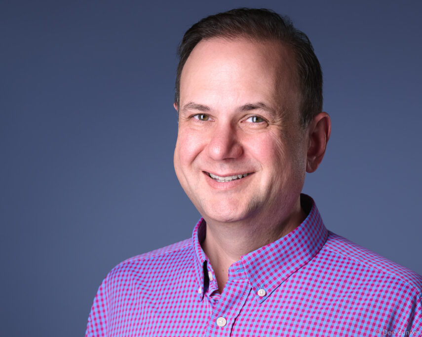 Male professional in pink gingham shirt smiles confidently against blue-gray studio backdrop in Houston corporate headshot session.