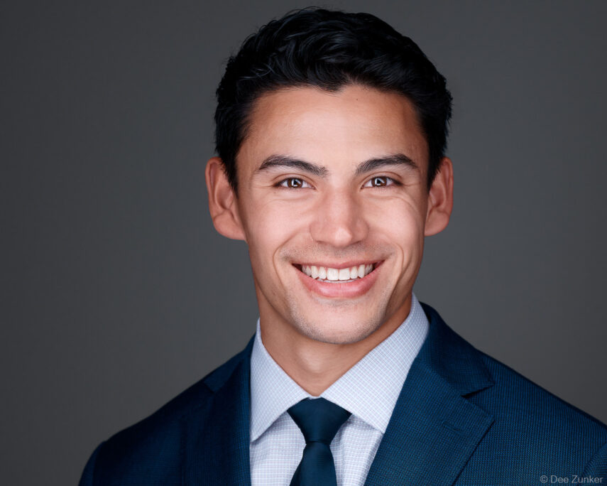 Smiling young male professional in navy suit and tie, studio corporate headshot against gray background, Houston TX.