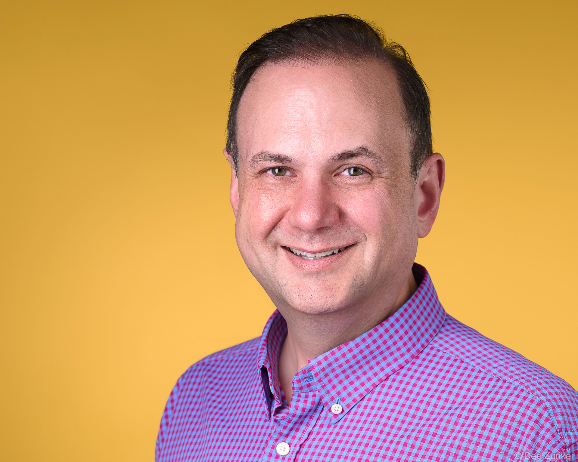 Smiling male professional in pink and blue checked shirt posed against a yellow background in a Houston corporate studio headshot.