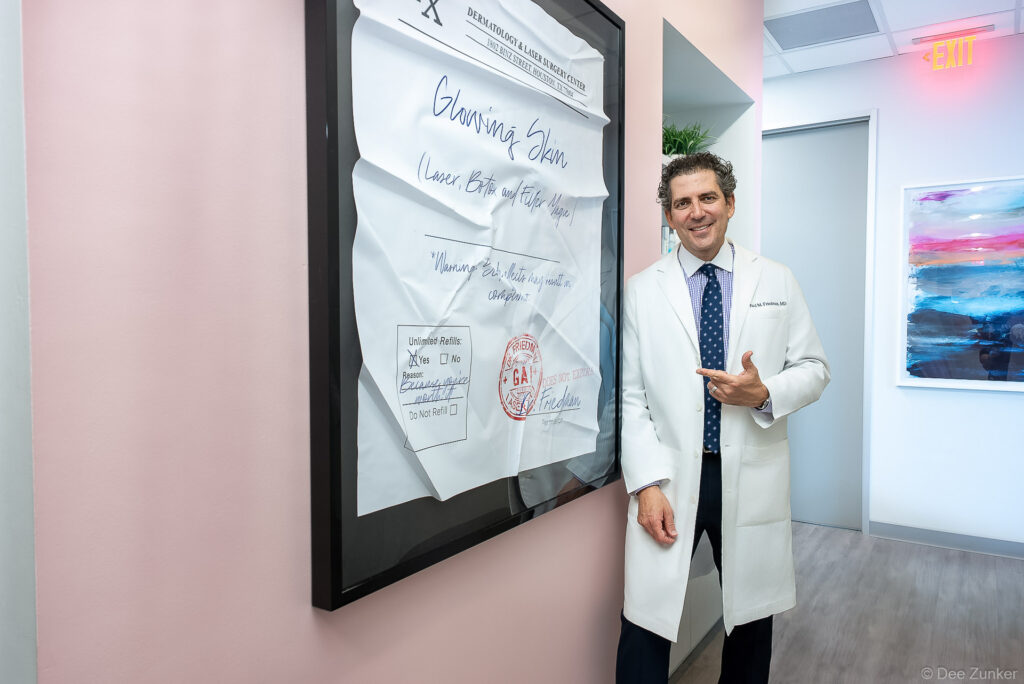 Dermatologist in white lab coat gestures beside framed oversized prescription artwork at Dermatology and Laser Surgery Center on Binz Street Houston.