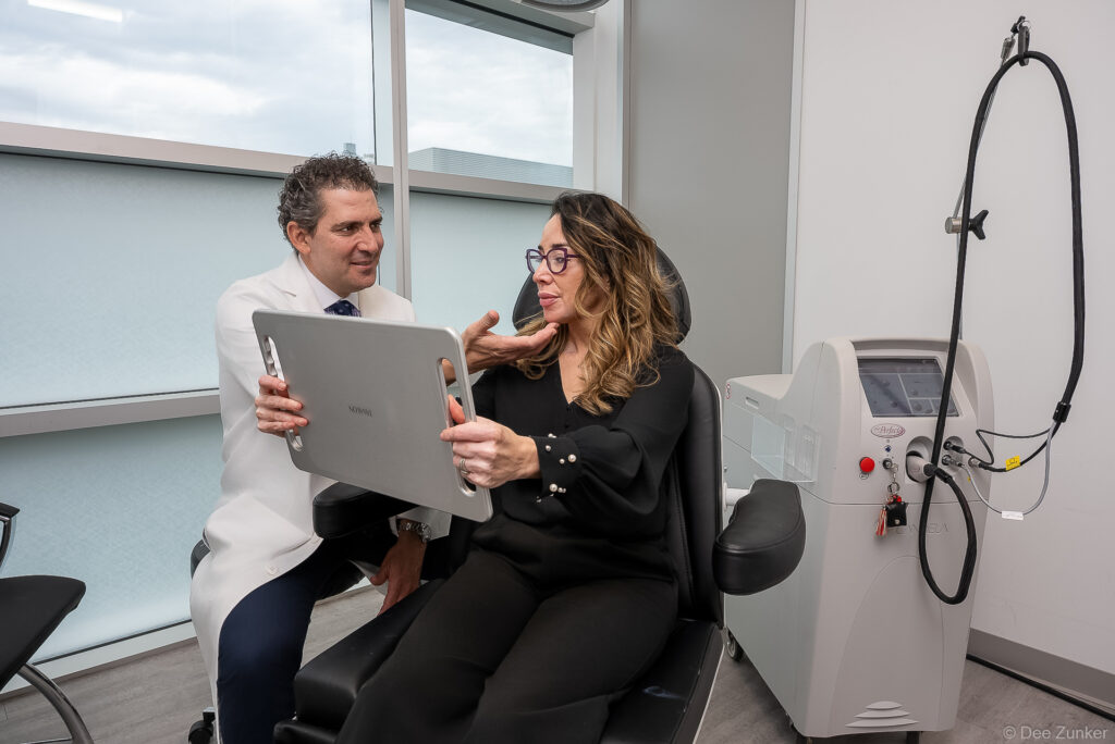 Dermatologist consults with female patient holding mirror at Houston Dermatology Laser Surgery Center on Binz, with laser equipment nearby.