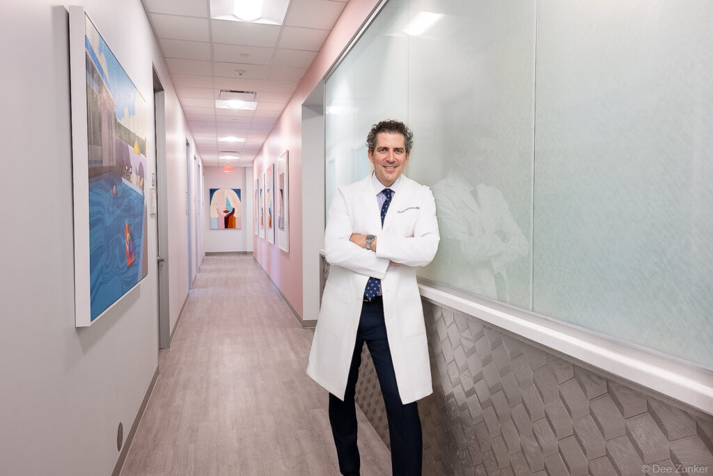 Houston physician in white coat stands arms crossed in a modern dermatology clinic hallway with colorful artwork, Binz location.