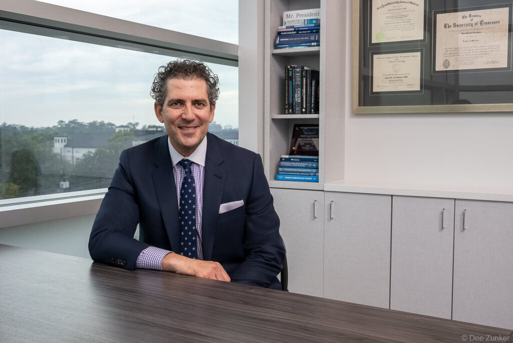 Male dermatologist in navy suit seated at desk with medical diplomas and textbooks, corporate headshot at Dermatology Laser Surgery Center Houston.