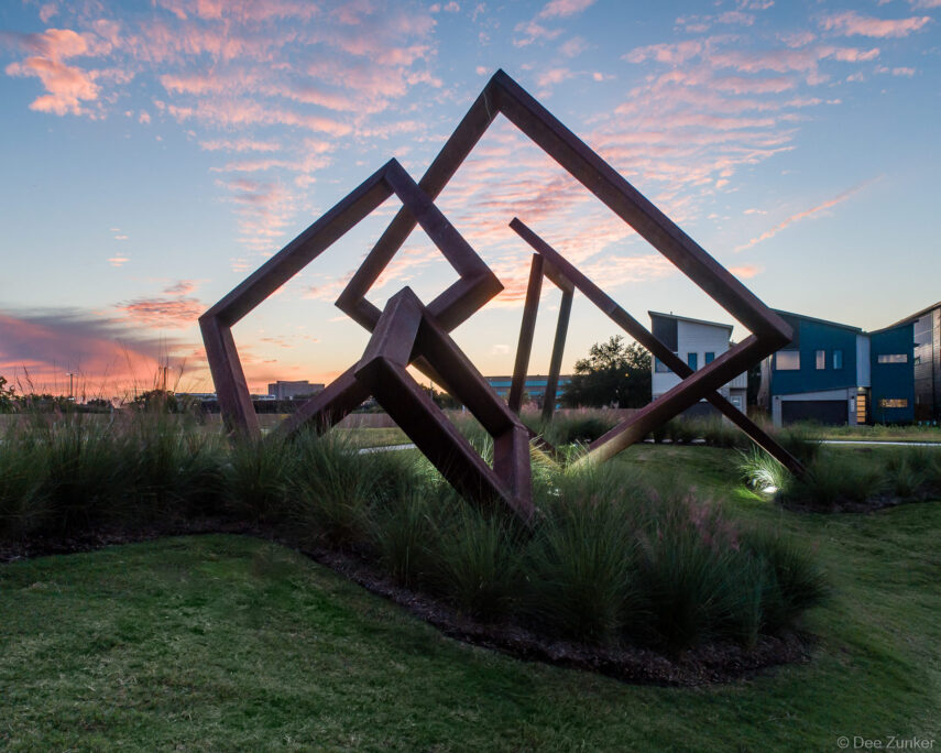 Aerial-perspective photo of large geometric steel cube sculpture at sunset, surrounded by ornamental grasses at a Houston property development.