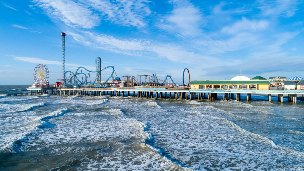 Aerial drone photo of the Pleasure Pier in Galveston, TX. The pier and rides stretch over the surf on a bright sunny morning.