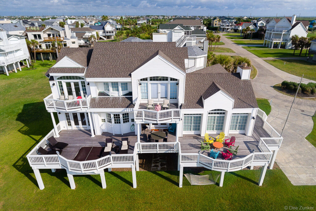 Drone aerial photo of large white multi-deck Galveston beach house with colorful Adirondack chairs and palm trees.