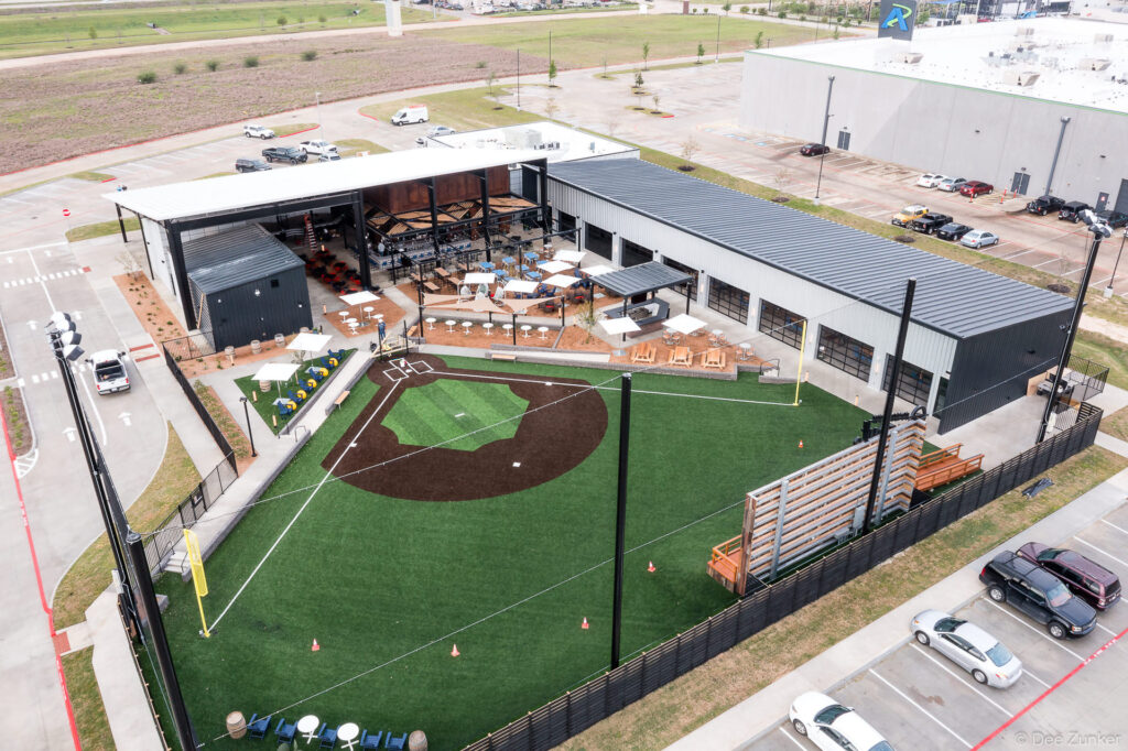 Aerial drone view of a baseball field and outdoor venue complex with artificial turf and open-air bar in Katy, TX.