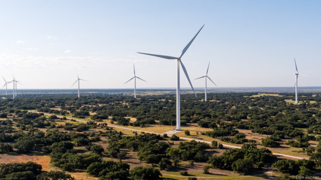 Aerial drone photo of Rattlesnake Wind Farm turbines rising above wooded West Texas landscape under clear blue sky.