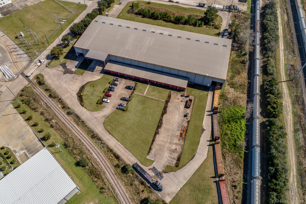 Aerial drone photo of a large steel warehouse facility beside railroad tracks in Houston TX, with parking lot and green grounds.