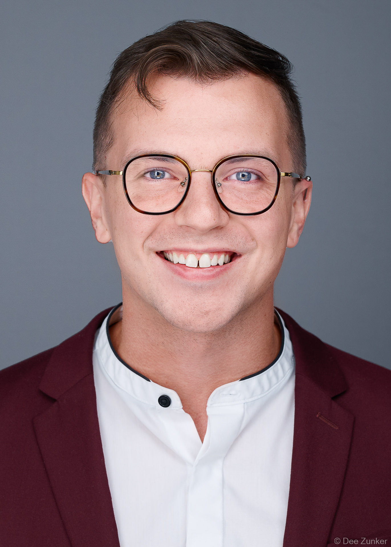 Male medical professional in burgundy blazer and glasses, ERAS residency application headshot, 5x7 format, photographed near Texas Medical Center, Houston TX with medium gray background.