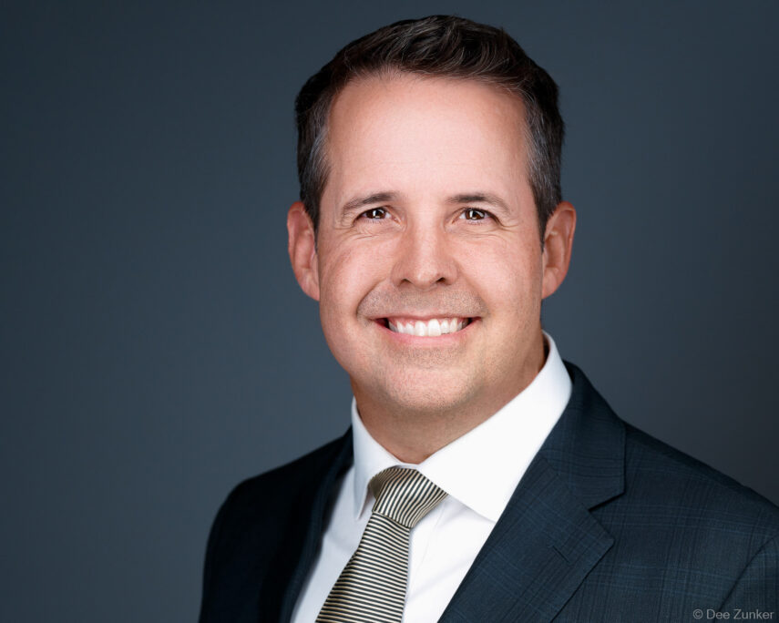 Male executive smiles in a professional studio headshot, wearing a dark suit and striped tie against a gray background in Houston.