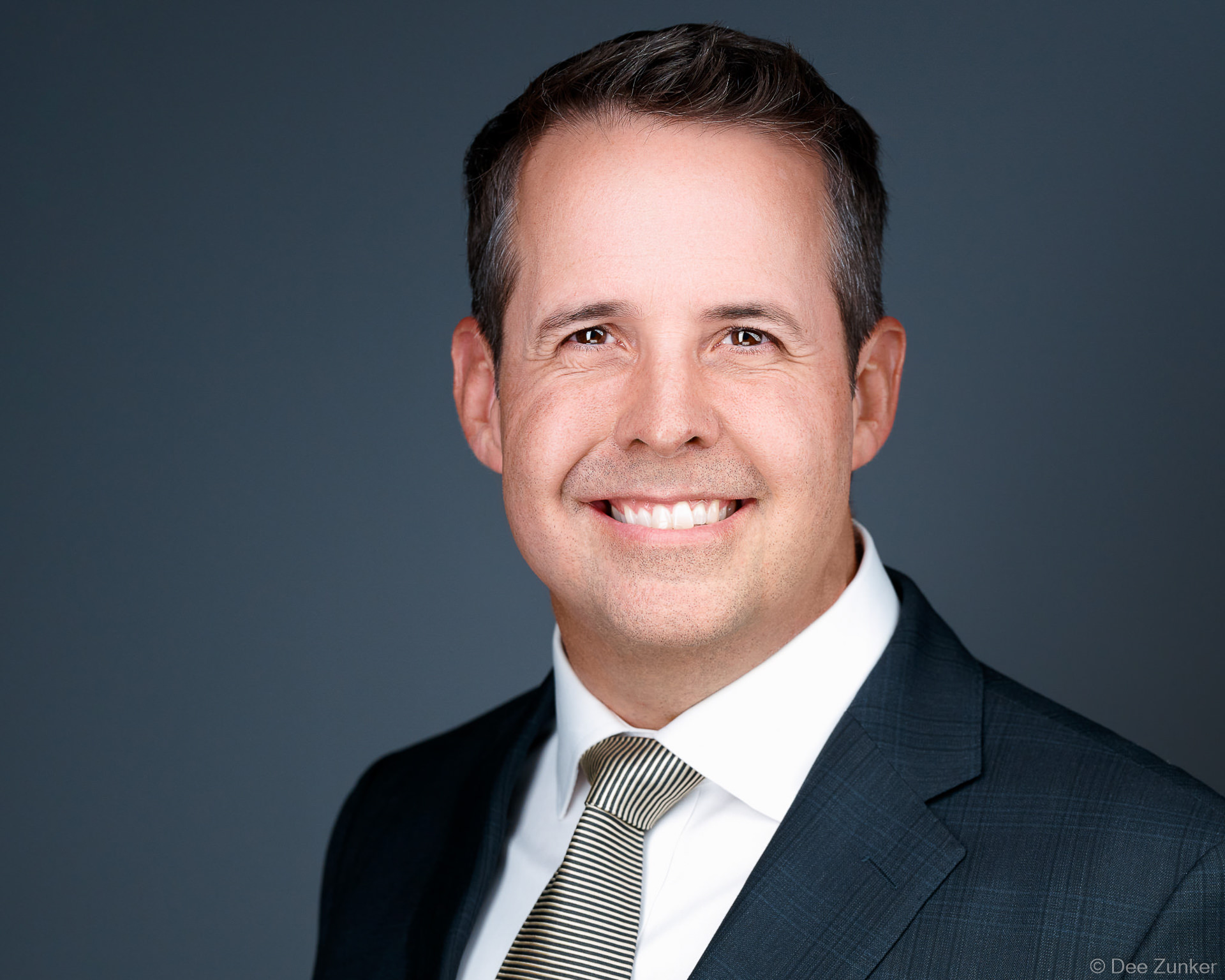 Male executive smiles in a professional studio headshot, wearing a dark suit and striped tie against a gray background in Houston.