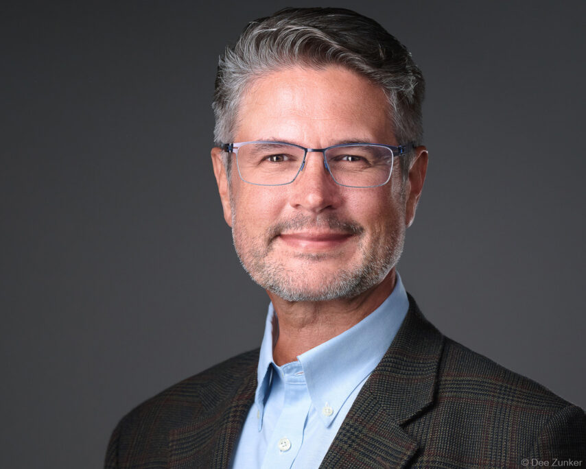 Professional male executive studio headshot, gray background, dark blazer and light blue shirt, glasses, Houston corporate photography.