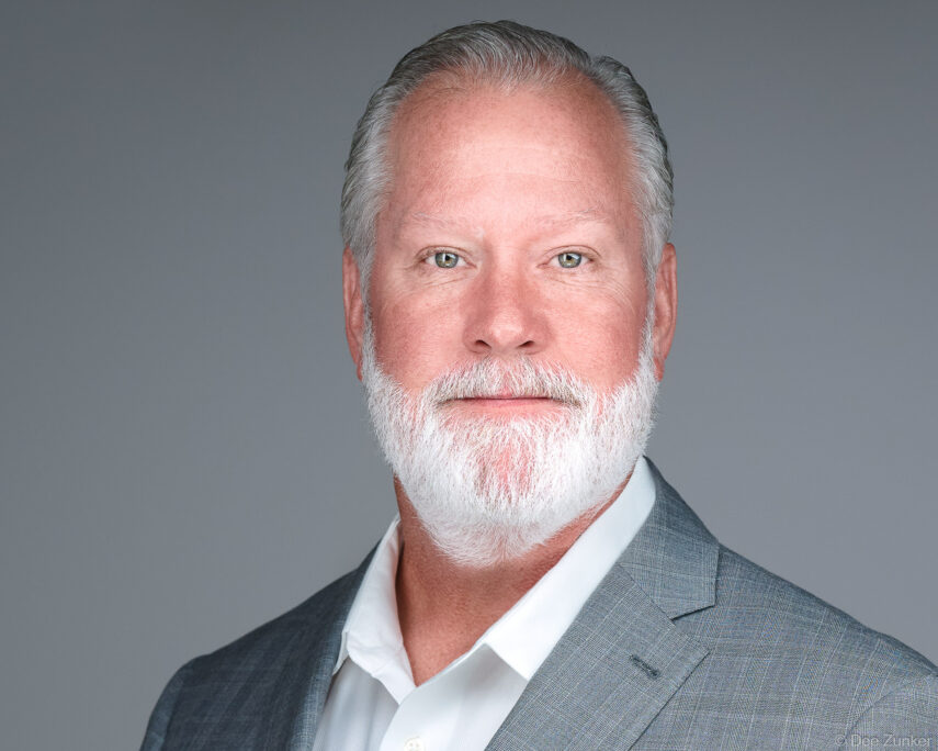 Professional corporate headshot of a senior male executive with white beard and gray blazer, shot on gray background in Houston studio.
