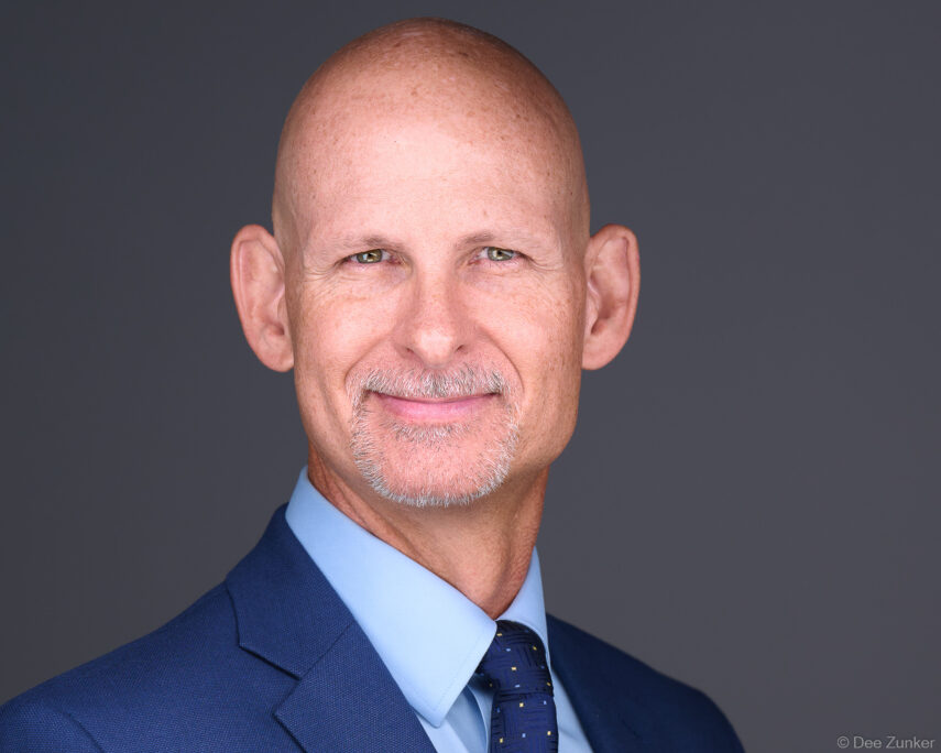 Bald male executive in navy suit and tie poses for a professional corporate headshot against a gray studio background in Houston.