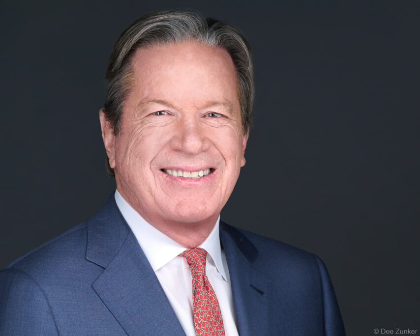 Senior male executive in navy suit and red tie smiles in a professional studio headshot against a dark gray background, Houston TX.