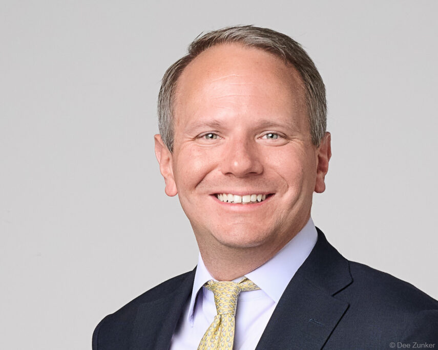 Male executive in navy suit and gold tie smiles in a professional studio headshot against a gray background, Houston, TX.