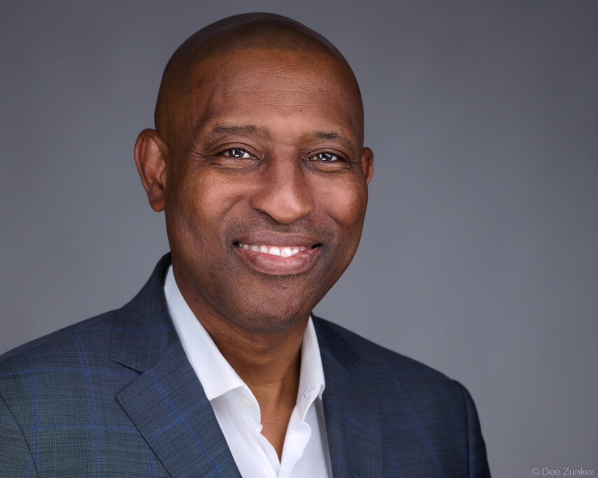 Smiling bald male executive in navy blazer and white shirt poses for a professional studio headshot against a gray background in Houston.