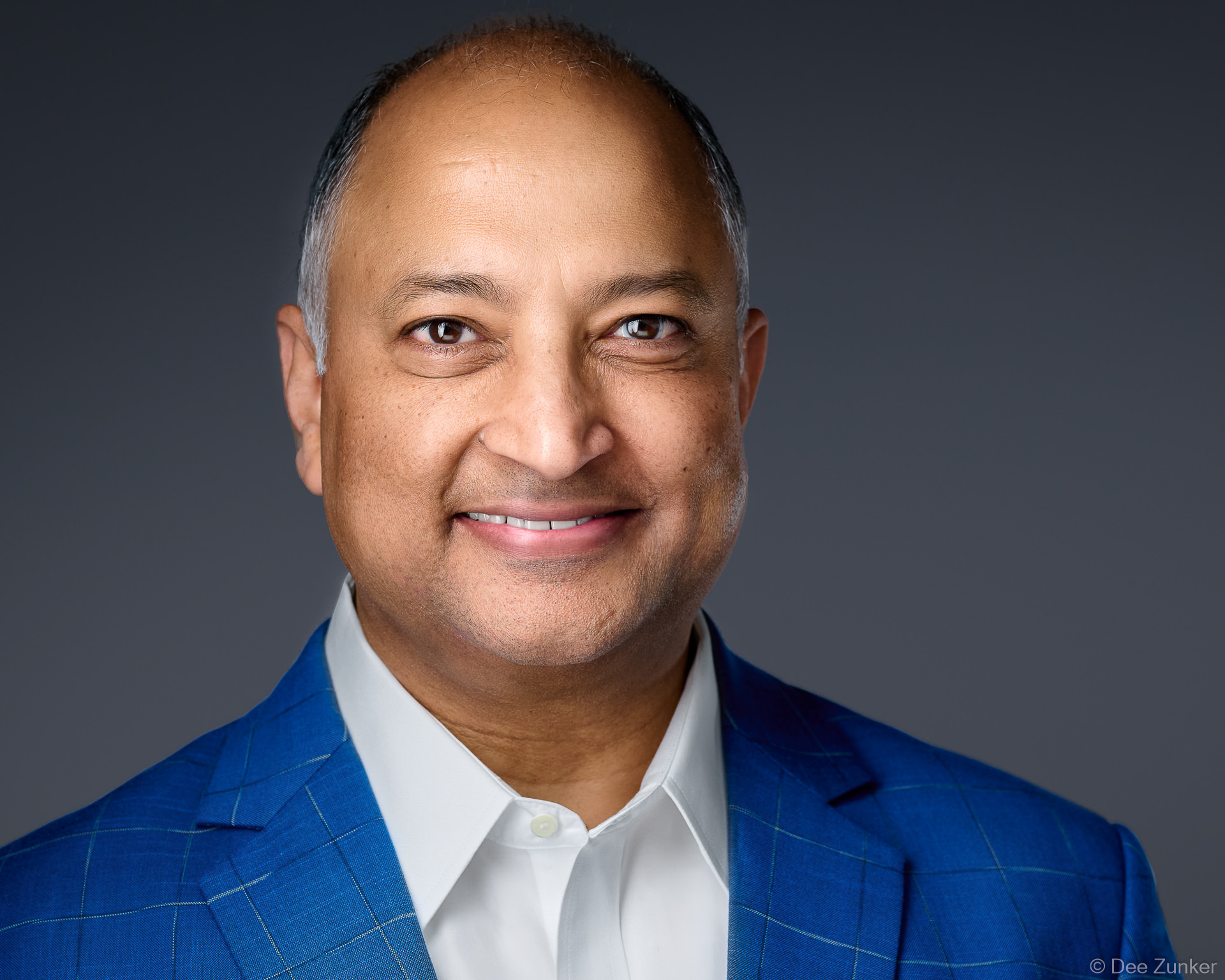 Male executive in blue plaid blazer smiles warmly against grey backdrop, professional studio headshot with soft 3-point lighting, Houston.