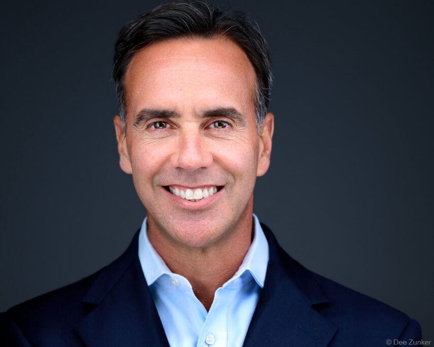 Male executive smiling in navy blazer and light blue shirt, professional studio headshot on dark grey backdrop, Bellaire Houston.